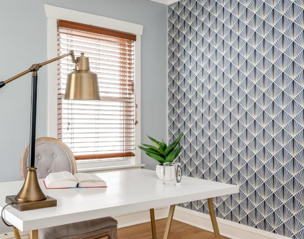 A home office featuring a geometric wallpaper design, a sleek white desk, and a brass table lamp.